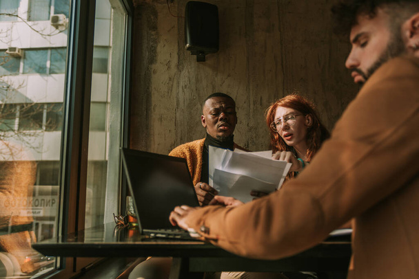 Low angle view van multiraciale mannelijke en vrouwelijke collega 's die samen papierwerk bespreken terwijl hun collega werkt aan lap top - Foto, afbeelding