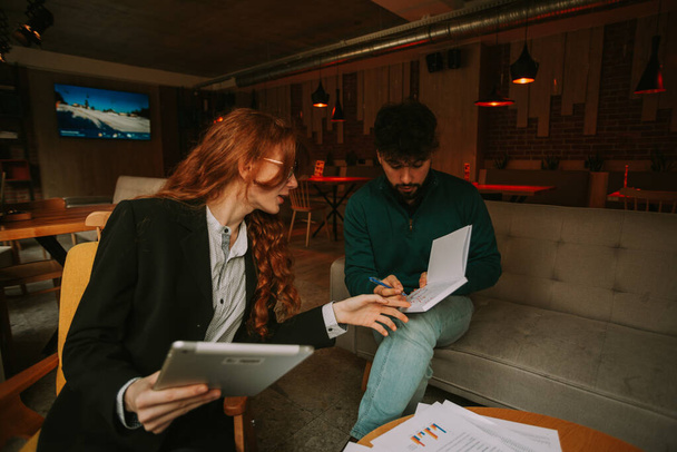 Handsome business person writing new ideas for the next project. His ginger female colleague is helping him. They are working at the coffee bar - Foto, imagen
