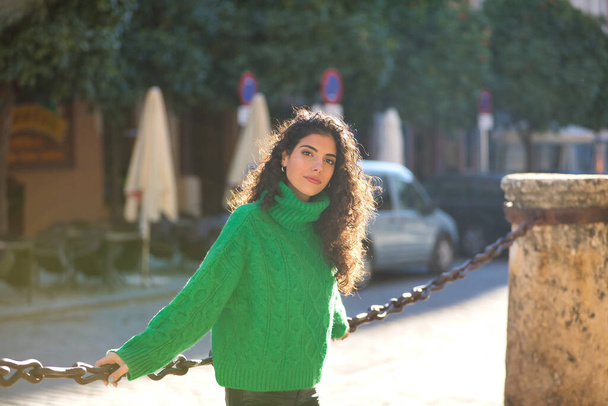 Young, beautiful, brunette woman with curly hair and a green turtleneck sweater, wrapped in the rays of the evening sun. Concept beauty, fashion, autumn, winter, cold, sun. - Photo, Image