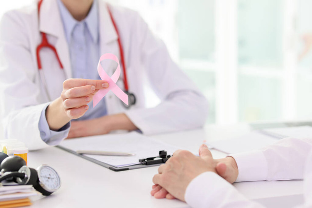 Doctor holds pink ribbon showing support for patient at appointment in clinic. Symbol of breast cancer awareness and struggle. Healthcare and treatment concept - Photo, Image