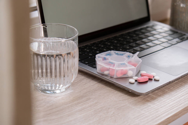 Organizer wekelijkse opnamen met glas water op de werkplek achtergrond en laptop Sluiten van de medische pil doos met doses tabletten voor dagelijks innemen van medicijnen met witte roze drugs en capsules. Geneesmiddelen - Foto, afbeelding