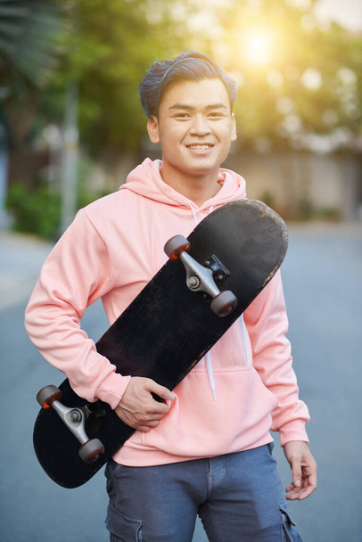Portrét usmívajícího se mladého muže držícího skateboard a usmívajícího se do kamery - Fotografie, Obrázek