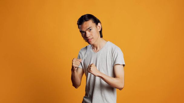 Asian man clenching fists and acting violent, causing conflict and being aggressive on camera. Young negative person being rebel and punching in studio, angry model on orange backdrop. - Photo, image