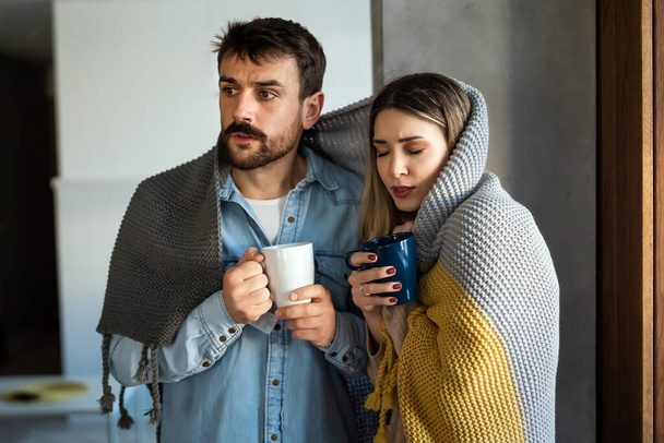 Sad sick young couple wrapped in blanket drinking hot tea trying to warm up. Freezing man and woman suffering from cold or flu fever or having trouble with central heating - Foto, imagen