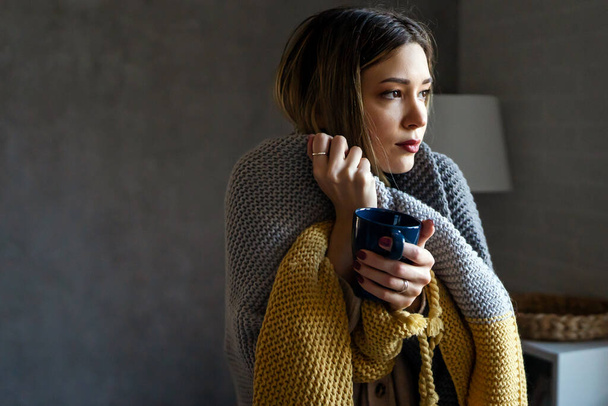 Sad sick young woman wrapped in blanket drinking hot tea trying to warm up. Freezing female suffering from cold or flu fever or having trouble with central heating - Foto, imagen
