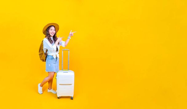Pretty Asian woman passenger in trendy fashion is pointing while carrying her luggage bag in happiness for travel and summer vacation isolated on yellow background with copy space - Photo, Image