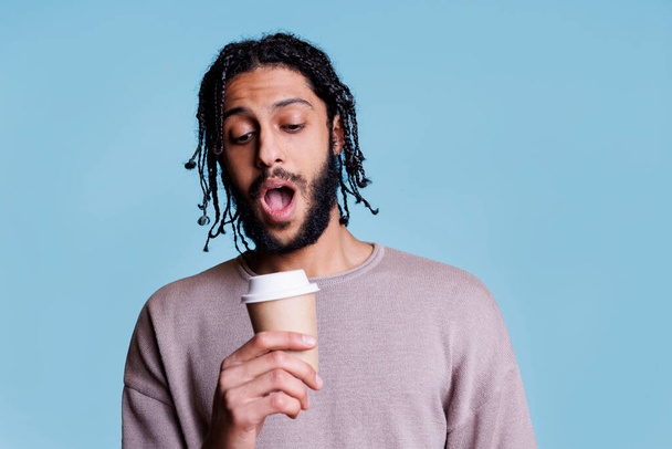 Surprised arab man holding coffee to go and looking at paper mug lid with shocked facial expression. Confused person drinking take out cappuccino cup while posing for studio shot - Foto, afbeelding