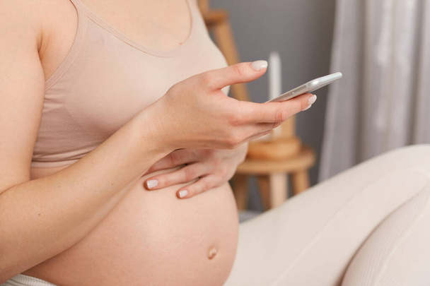 Tanımlanamayan hamile bir kadın, bej bir bluzla yatakta oturuyor. Cep telefonu kullanıyor, mesajlar yazıyor, internette geziniyor, elinde akıllı bir telefon var.. - Fotoğraf, Görsel