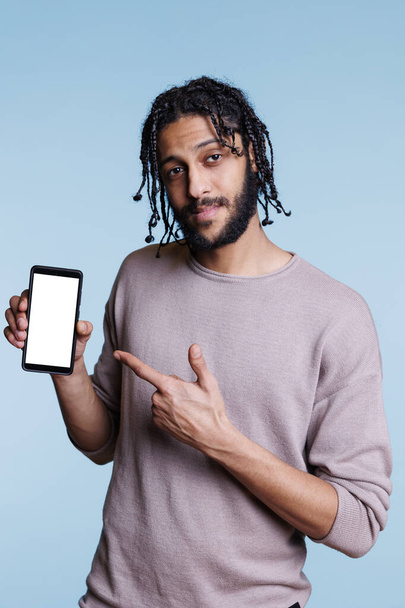 Pewny siebie arabski reklamodawca trzymający smartfona i wskazujący biały pusty ekran dotykowy patrząc w kamerę. Mężczyzna prezentujący telefon komórkowy z pustym wyświetlaczem, produkt reklamowy - Zdjęcie, obraz