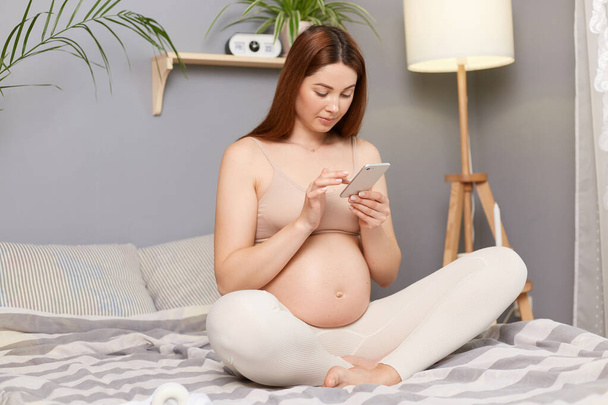 Beautiful pregnant brown haired caucasian woman sitting in bed in lotus pose using mobile phone reading new in social networks typing messages browsing internet. - Fotoğraf, Görsel
