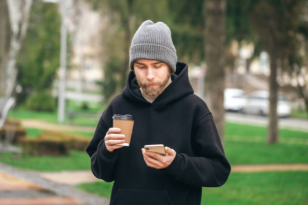 Ένας νεαρός άνδρας με γενειάδα και καπέλο χρησιμοποιεί ένα smartphone και πίνει καφέ από ένα χάρτινο κύπελλο στο πάρκο, θολό φόντο. - Φωτογραφία, εικόνα