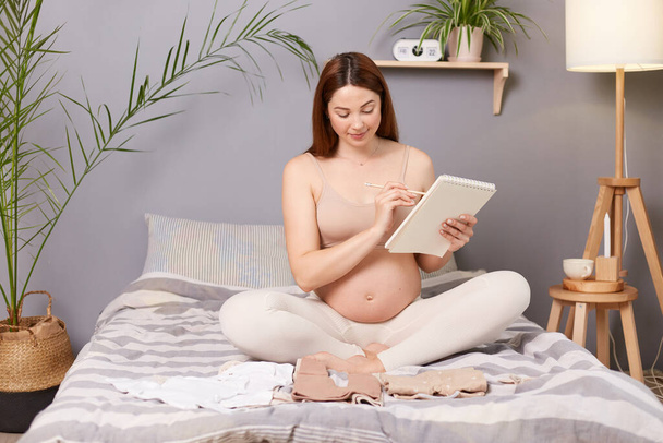 Smiling happy pregnant brown haired caucasian woman sitting in bed expecting baby holding paper notebook planning preparing for giving birth. - Foto, afbeelding