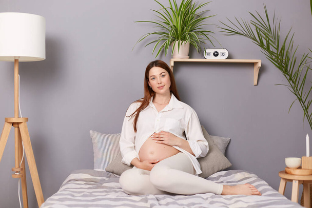 Adorable calm pregnant brown haired caucasian woman sitting in bed resting embracing her belly looking at camera resting at home on last months of pregnancy. - Fotó, kép