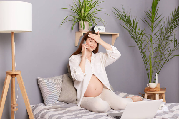 Pregnancy health. Phone consultation. Unhealthy sick pregnant brown haired caucasian woman sitting in bed and talking on smart phone with doctor having headache and high temperature. - Fotoğraf, Görsel