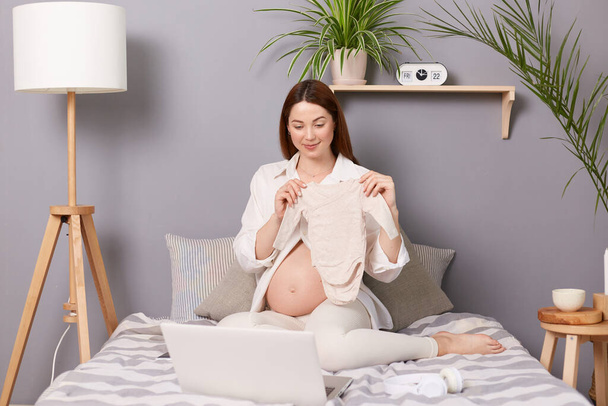 Smiling attractive pregnant brown haired caucasian woman sitting in bed having video call on laptop online conversation showing new little baby's clothing to notebook camera. - Photo, image