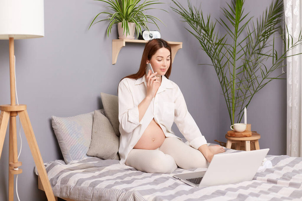 Busy pregnant brown haired caucasian woman sitting in bed working on laptop and talking on smart phone, combines maternity leave on last months of pregnancy and online work. - Photo, Image