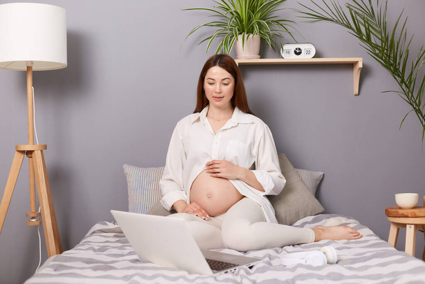Concentrated pregnant brown haired caucasian woman sitting in bed in front of laptop watching movie spending leisure time at home resting. - Photo, Image