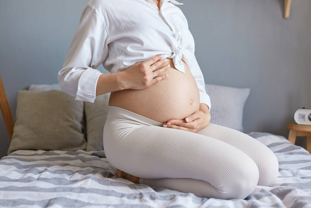 Een gezonde zwangerschap. Verwacht een bevalling. Onherkenbare onbekende zwangere vrouw draagt witte kleding zittend in bed in lichte slaapkamer met haar blote buik. - Foto, afbeelding