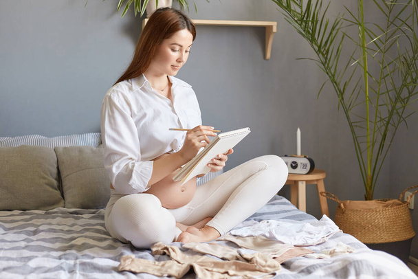 Serious responsible pregnant woman wearing white clothing sitting in bed with paper notebook making list planning collects child's things in the maternity hospital. - Фото, изображение