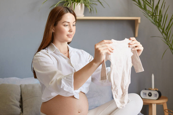 Satisfied caucasian young adult pregnant woman with bare belly wearing white clothing sitting in bed in light bedroom, future mother holding little kid's bodysuit admiring tiny child'd clothes. - Foto, Imagen