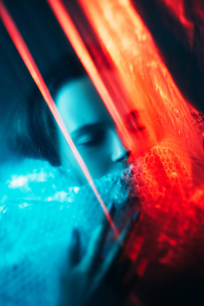 Ghost spirit. Soul silence. Dream mystery. Red blue color light defocused tranquil cold woman face with closed eyes wrapped in polyethylene plastic film on dark. - Фото, изображение