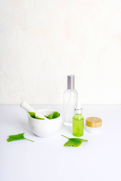 Mortar and pestle with fresh green ginkgo leaves, cosmetic serum and tonic bottles, cream jar on white background. Natural handmade cosmetics concept. - Valokuva, kuva