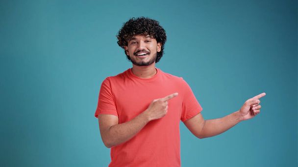 Smiling young handsome indian man wearing casual clothes on a blue studio background pointing finger at blank space - 写真・画像
