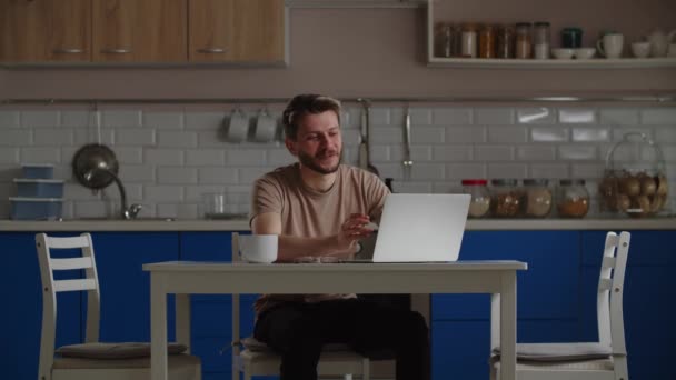 Uśmiechnięty mężczyzna siedzący przy stole z filiżanką kawy i laptopem komunikuje się z kimś przez wideo połączenie. Męski wolny strzelec pracujący w domu omawia szczegóły nowego projektu pracy ze swoim - Materiał filmowy, wideo