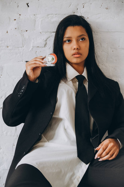 A dark-skinned young woman sits pretentiously in a white room and eats sushi with chopsticks. African American woman in a business suit eats rolls. - Foto, Imagen