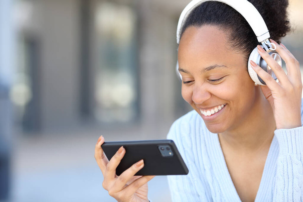 Happy black woman wearing headphone watching videos on horizontal smart phone in the street - Фото, зображення