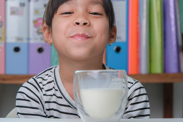 Nettes asiatisches Mädchen trinkt zu Hause im Wohnzimmer ein Glas Milch. Kleines Mädchen, das morgens vor dem Schulbesuch Milch trinkt. Gesunde Ernährung in der Kindheit. - Foto, Bild