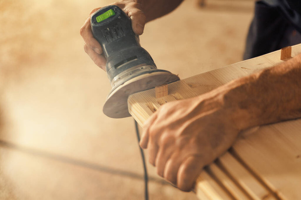 Las manos del carpintero agarran una lijadora. El artesano de madera, un profesional experimentado, sostiene una tabla o pieza de madera con manos fuertes y precisas, lijándola cuidadosamente con una máquina que vibra arena - Foto, imagen