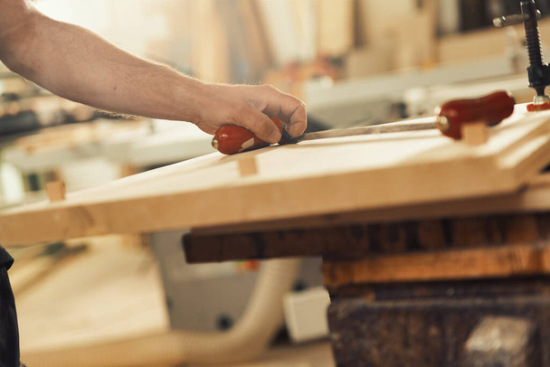 n ένα πριονιστήριο ή εργαστήριο ξυλουργικής βιοτεχνίας, το χέρι ενός ξυλουργού πιάνει τη λαβή ενός μαχαιριού που στηρίζεται σε ανοιχτόχρωμο ξύλο. Ο τεχνίτης έχει δυνατά και ανδρικά χέρια. - Φωτογραφία, εικόνα