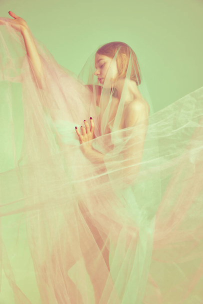 Young adorable woman with slim body shape standing alone covered transparent fabric over pastel color palette background. Natural beauty of female body concept. Highlights effect. - Φωτογραφία, εικόνα