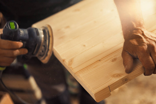 Τα χέρια του ξυλουργού πιάνουν ένα τριβείο. Ο ξυλουργός, έμπειρος επαγγελματίας, κρατά μια σανίδα ή ένα κομμάτι ξύλο με δυνατά και ακριβή χέρια, τρίβοντάς το προσεκτικά με μια μηχανή που δονείται άμμος - Φωτογραφία, εικόνα