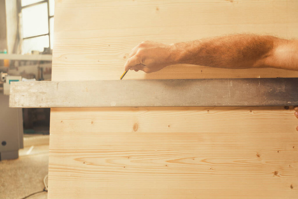A háttérben egy műhely, előtér egy nagy fa felület és egy négyzet. Carpenter karja kinyújtja egy ceruzával, hogy jelölje meg a méréseket, kettéosztva a keretet, így bőséges COPYSPACE - Fotó, kép