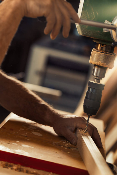 Carpenter uses drill press in his workshop. With his left hand, taking calculated risks, he holds and guides the wood, while lowering the lever with his right hand to control the exact spot and pressu - Photo, image