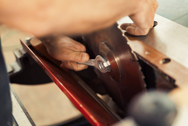 Στιγμιότυπο ενός κλειδιού που σφίγγει ένα παξιμάδι για να εξασφαλίσει μια κυκλική λεπίδα πριονιού. Η άποψη στο επίκεντρο περιβάλλεται από επιλεκτικές θολές των χεριών του ξυλουργού κατά την εργασία και τα μηχανήματα κοπής στο backgrou - Φωτογραφία, εικόνα