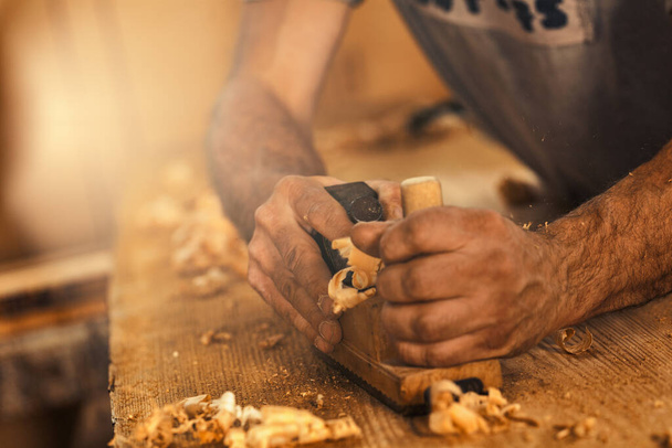 The wood artisan, a robust and strong man known as a carpenter, hand planes a board in the traditional manner, producing shavings of sawdust that look like golden curls of wood. It is a strenuous and  - Photo, Image