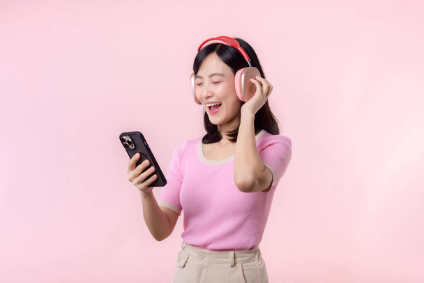 若い陽気なアジアの女性の肖像画は、ピンクに対するスマートフォンの音楽アプリケーションによってオーディオを聞いてお楽しみください。ヘッドフォンで幸せな笑顔の女性の人。音、レジャー、ライフスタイル、テクノロジーの概念 - 写真・画像