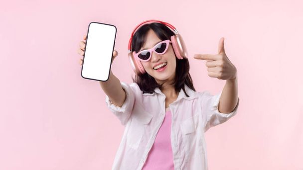Glücklich lächelnde fröhliche asiatische Frau mit drahtlosen Kopfhörern, die leere Bildschirme zeigen Mobiltelefon oder neue Smartphone-Musik-Anwendung Werbung Attrappe isoliert auf rosa Studiohintergrund. - Foto, Bild