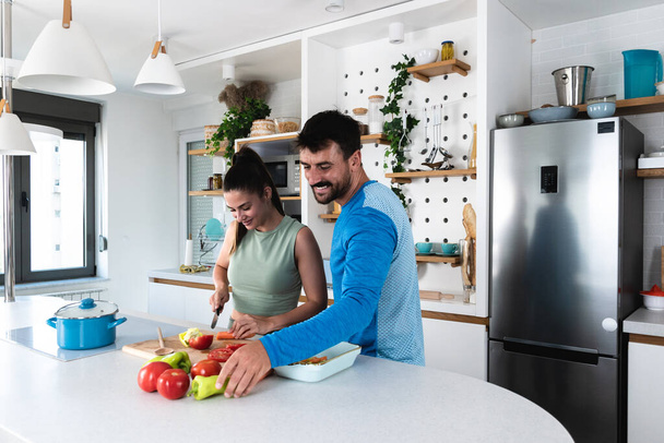 幸せな若いカップルは健康的な有機食品を準備しながらキッチンで楽しみを持っています。家庭のキッチンでベジタリアン料理をしながら、美しいスポーツの人々が話していると笑顔. - 写真・画像