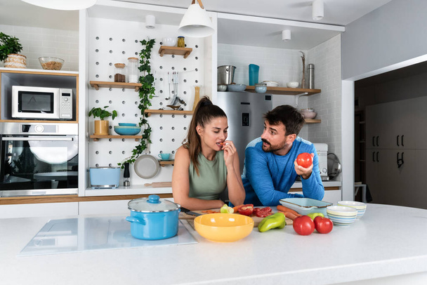 Happy young couple have fun in kitchen while preparing healthy organic food. Beautiful sports people are talking and smiling while cooking vegetarian meal in domestic kitchen at home. - 写真・画像
