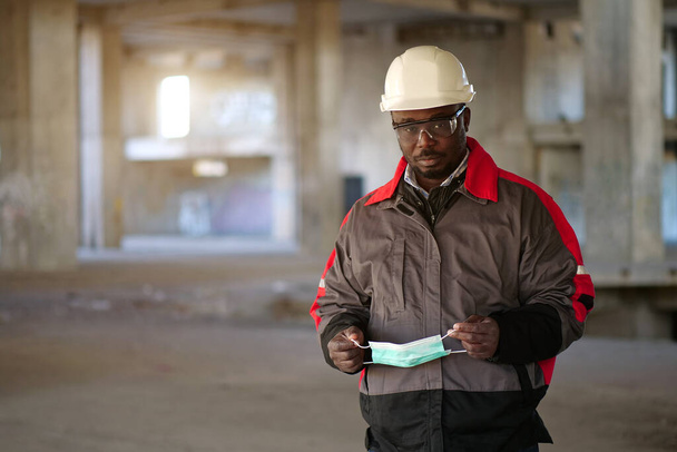 Αφροαμερικάνος κατασκευαστής στέκεται στο εργοτάξιο, κοιτάζει την κάμερα και κρατά στα χέρια του ιατρική προστατευτική μάσκα - Φωτογραφία, εικόνα