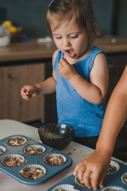 かわいい赤ちゃんの女の子は、ベーキングカップにマフィン打者を振りかけ、自宅で焼きながらチョコレートを食べます。幸せな家庭の子供の概念. - 写真・画像