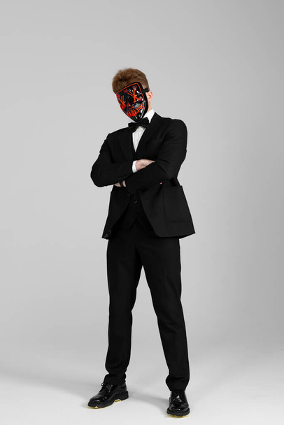 Stylish young man in a sharp black formal suit wearing a bright face mask. Concealed identity and personality. Hidden intentions. Identity substitution. Isolated on white background. - 写真・画像