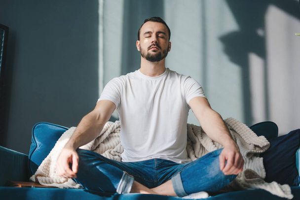 Calm satisfied guy in stylish casual clothes sitting on a sofa in a living room with closed eyes, meditates in the lotus position. Meditation, relaxation, rest - Foto, afbeelding