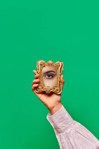 Main féminine tenant petit miroir avec reflet de charmant oeil de femme avec un maquillage lumineux sur fond vert. Mode, beauté, créativité, publicité. Émotions humaines, concept d'expression - Photo, image