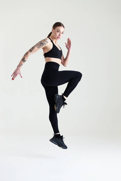 A young girl with brown hair, with a tattoo in a black top and leggings on a white background. Sports, fitness, jumping - Fotó, kép