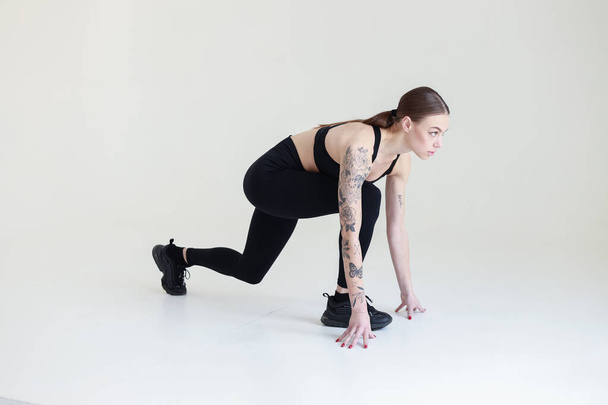 A young girl with brown hair, with a tattoo in a black top and leggings on a white background. Sports, fitness, starts running - Foto, imagen